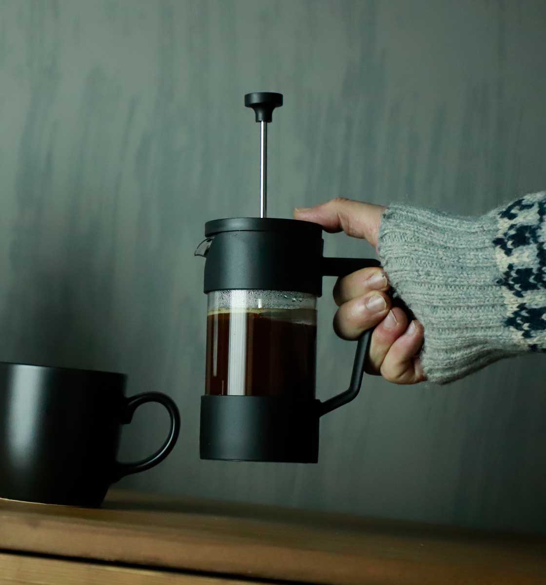 Ahşap tezgahın üzerindeki kupanın yanında hava tutulan içi kahve dolu bir french press.