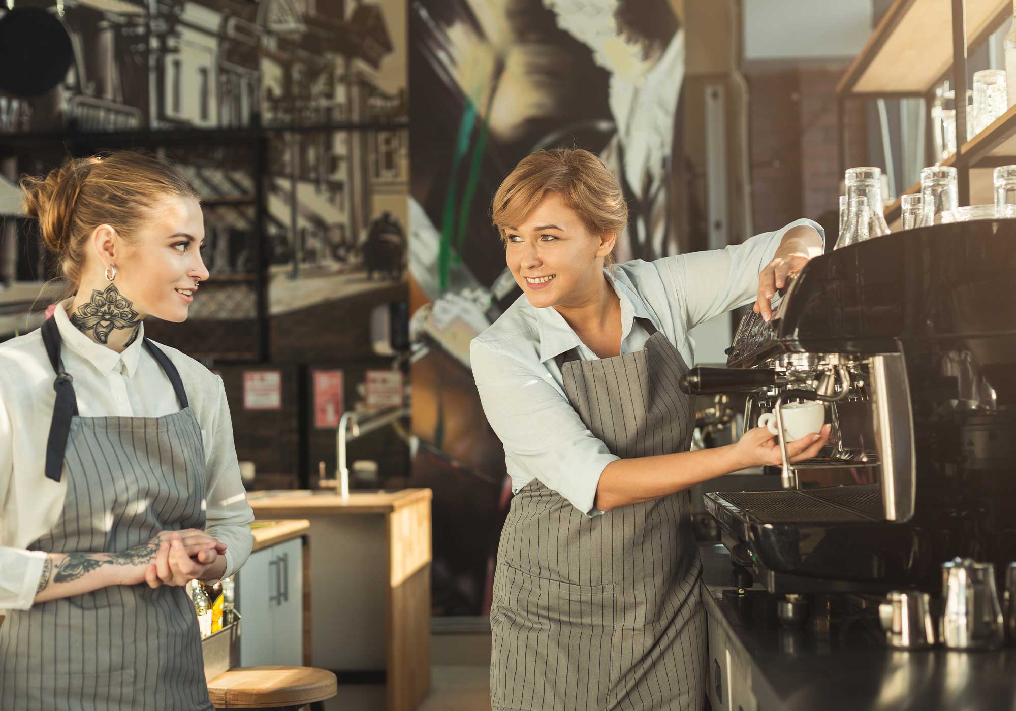 Bir kafe ortamında sohbet eden iki kadın barista yan yana yer alırken, sağ taraftaki espresso makinesi ile kahve demlemektedir.