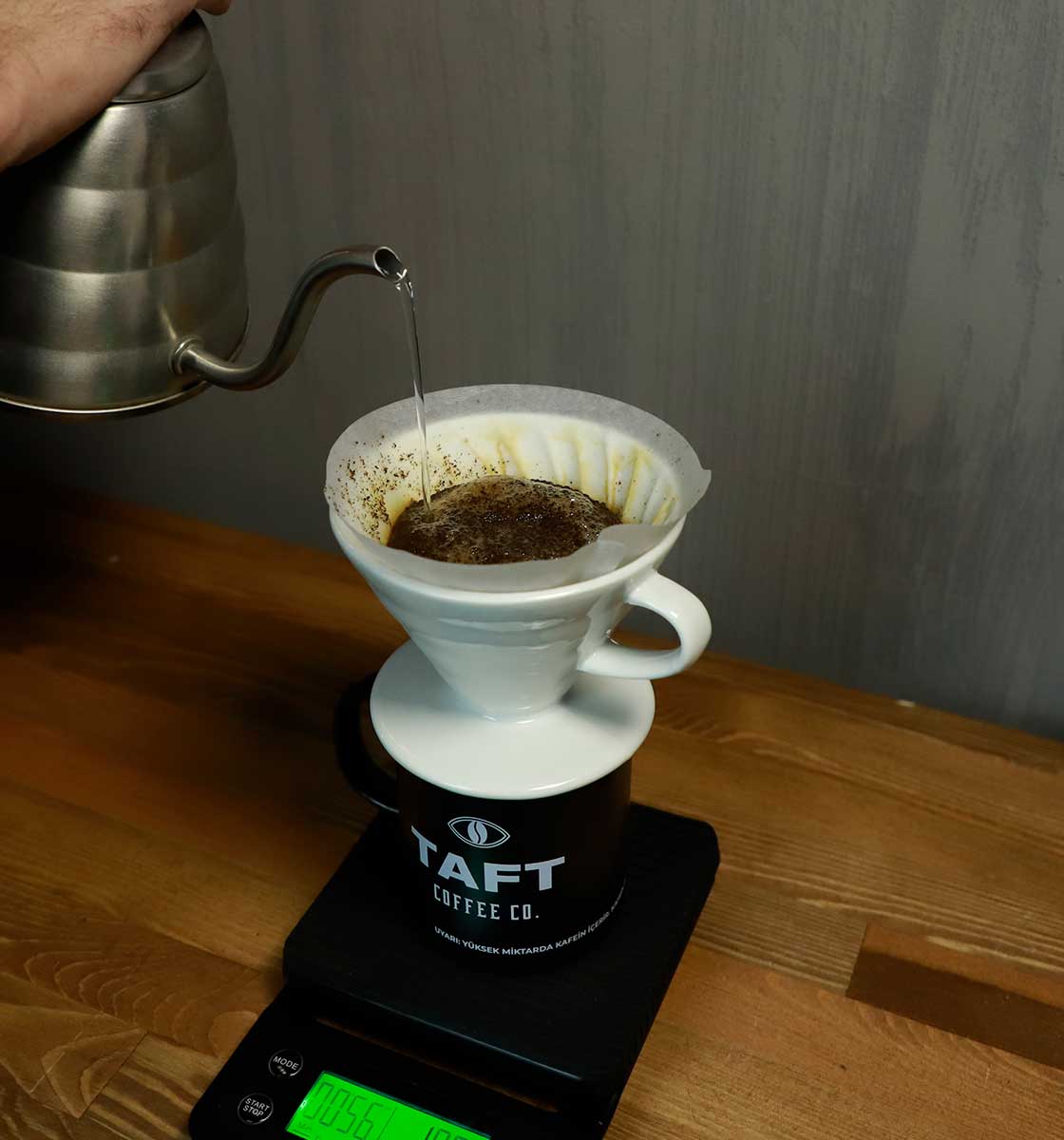 Ahşap tezgah üzerindeki hassas tartınız üzerinde yer alan TAFT Kupa içerisinde v60 demleme ekipmanıyla filtre kahve demlenerek aktarılıyor.